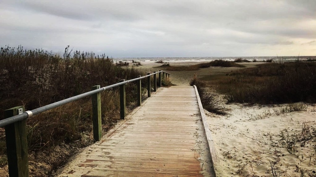 A gloomy overcast day walking down a sandy wood lined boardwalk. Darkened dunes and tall grasses line the walkway. In the distance waves crash against the beach.