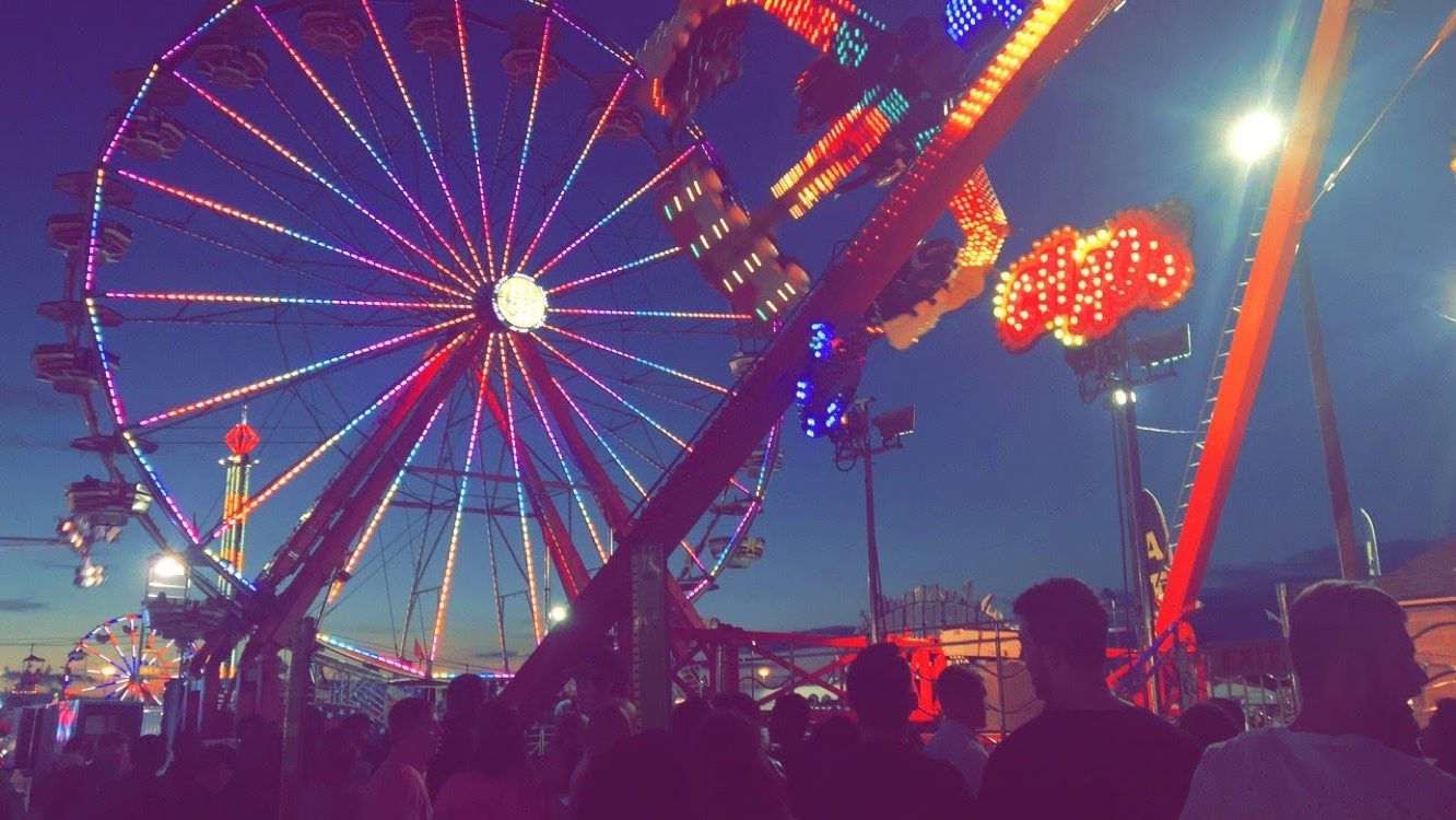 A vibrant blue night sky with illuminated fair rides taking up the space. A Ferris wheel twinkles on the left with a rotating spinning ride swinging on the left. 