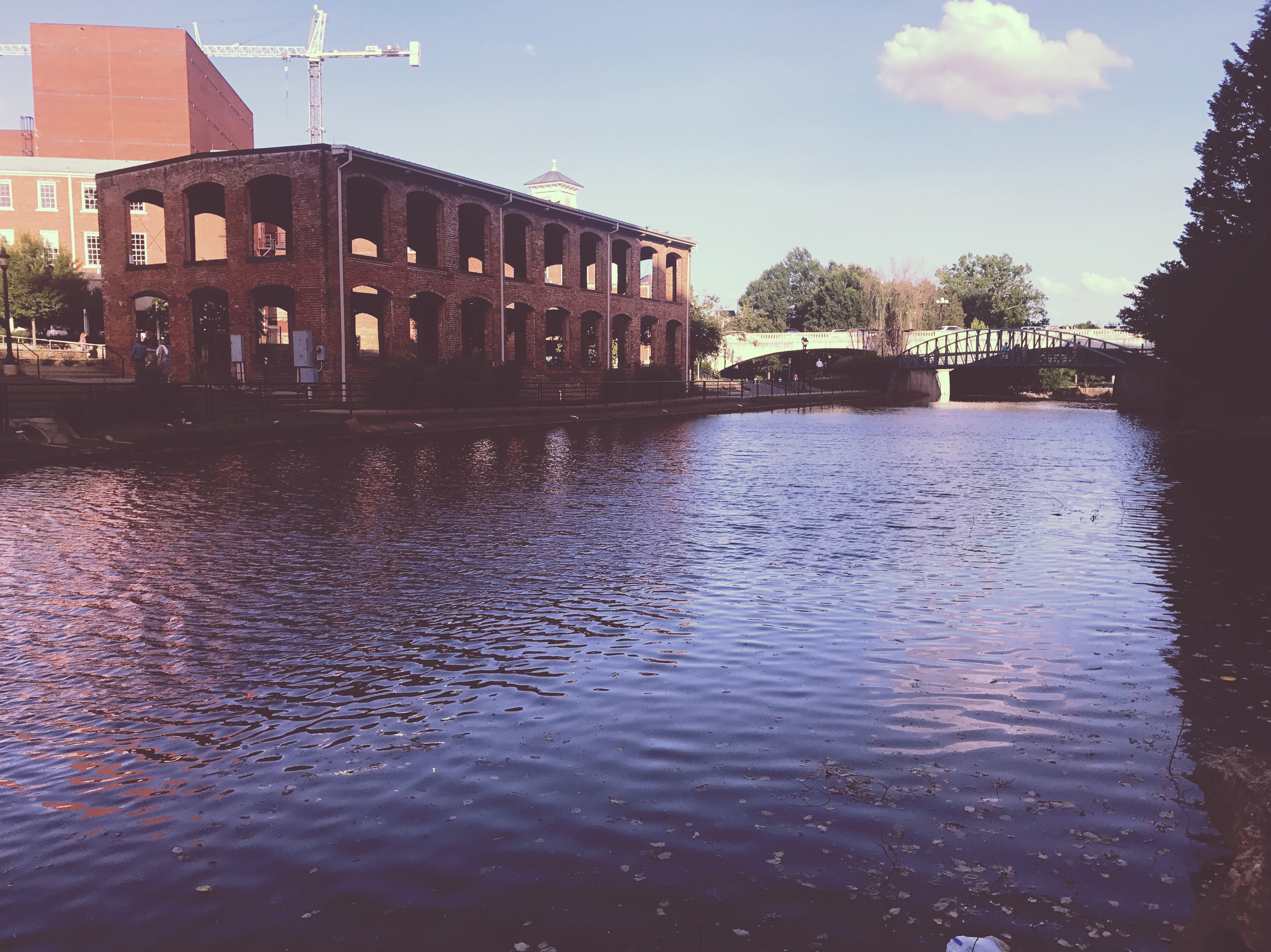 In a warm pink glow the Reedy River moves softly through Greenville. A large brick building with large arched windows sits across the river near the bridge. 