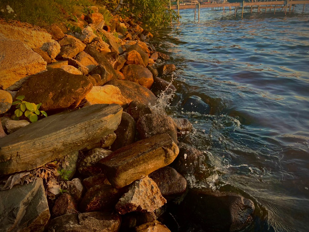 A longer line of rocks some more jagged line the other side of the lake. The deep blue water splashes against them with a tall plume of droplets dancing up in the center where the wave has crashed.