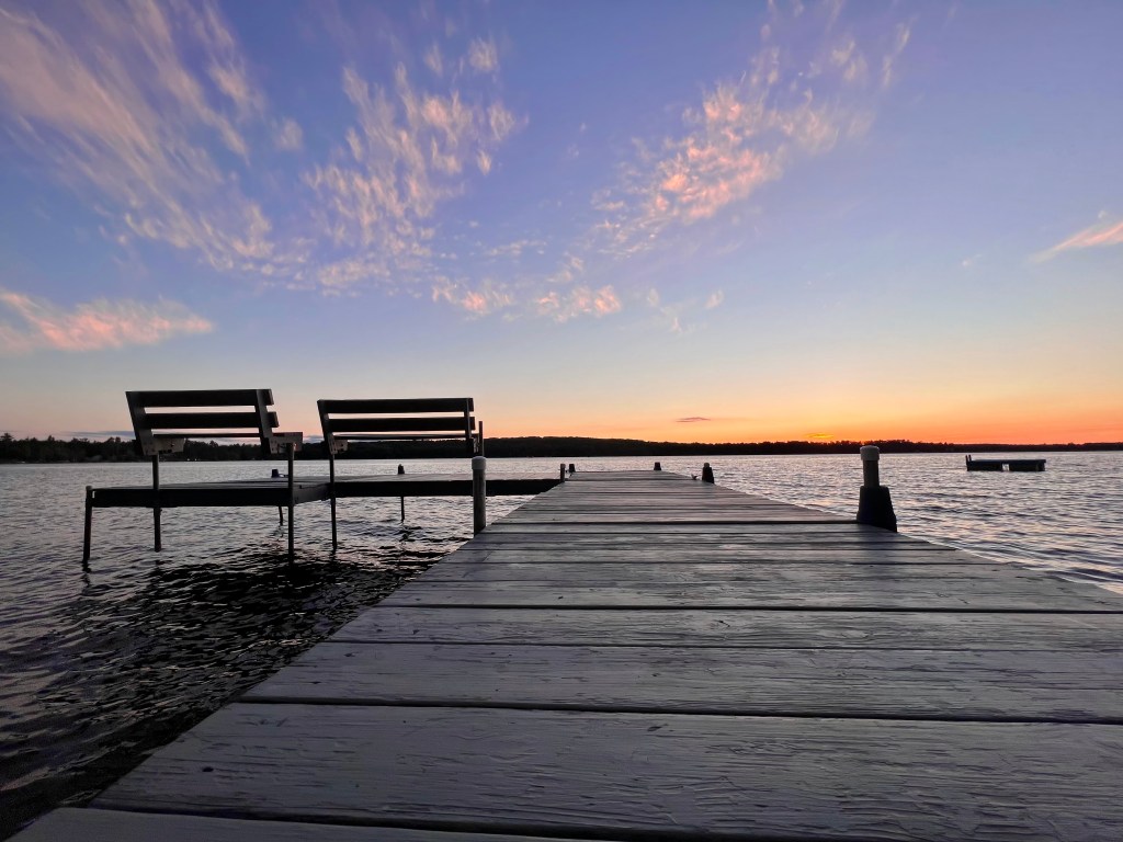 From the dock, it extends outwards from a low perspective. The two benches at the end of the dock sit next to each other on the left like good friends. The orange and pink sunset fading in the distance