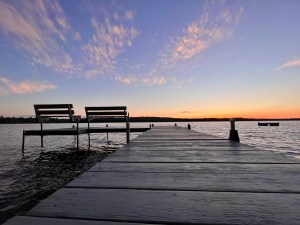 From the dock, it extends outwards from a low perspective. The two benches at the end of the dock sit next to each other on the left like good friends. The orange and pink sunset fading in the distance