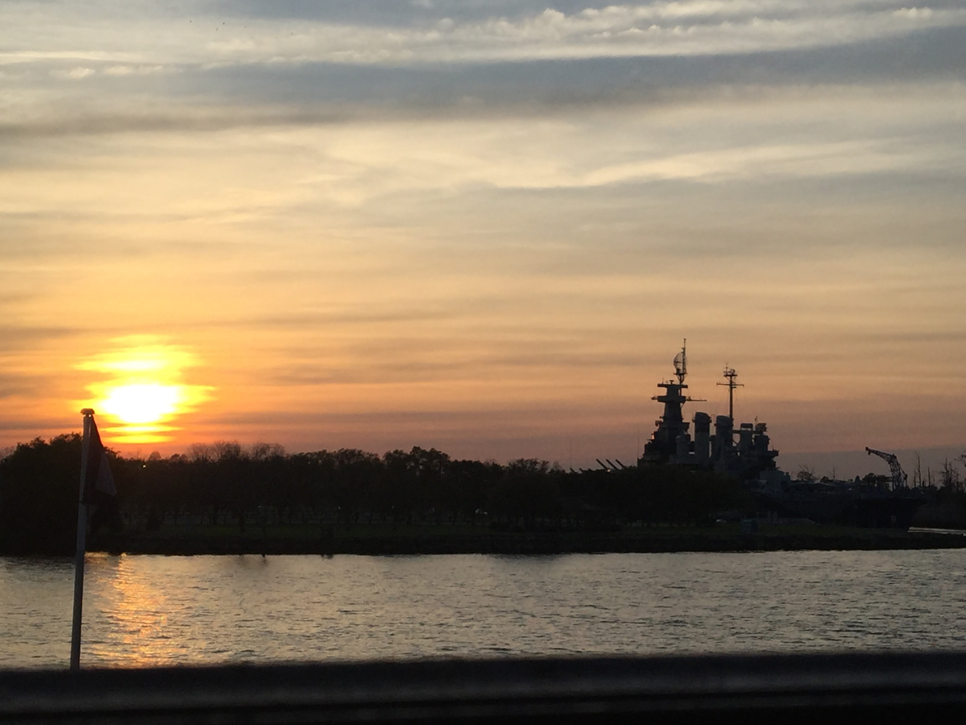 Looking across the Cape Fear River as the sun sets in pastel orange hues, the docked military ship creates a silhouette with the trees. 