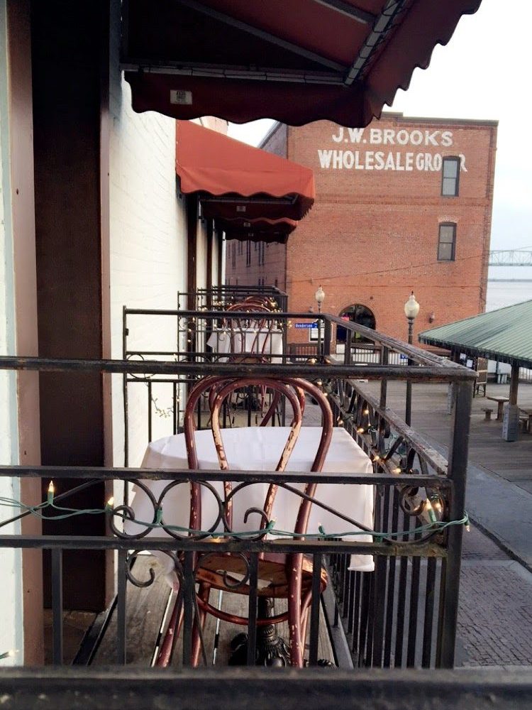 From an iron balcony looking along a white building with red awnings you see other small balconies lined perfectly with white table cloths and antique wooden chairs. The red brick brooks factory to the right in the distance. 