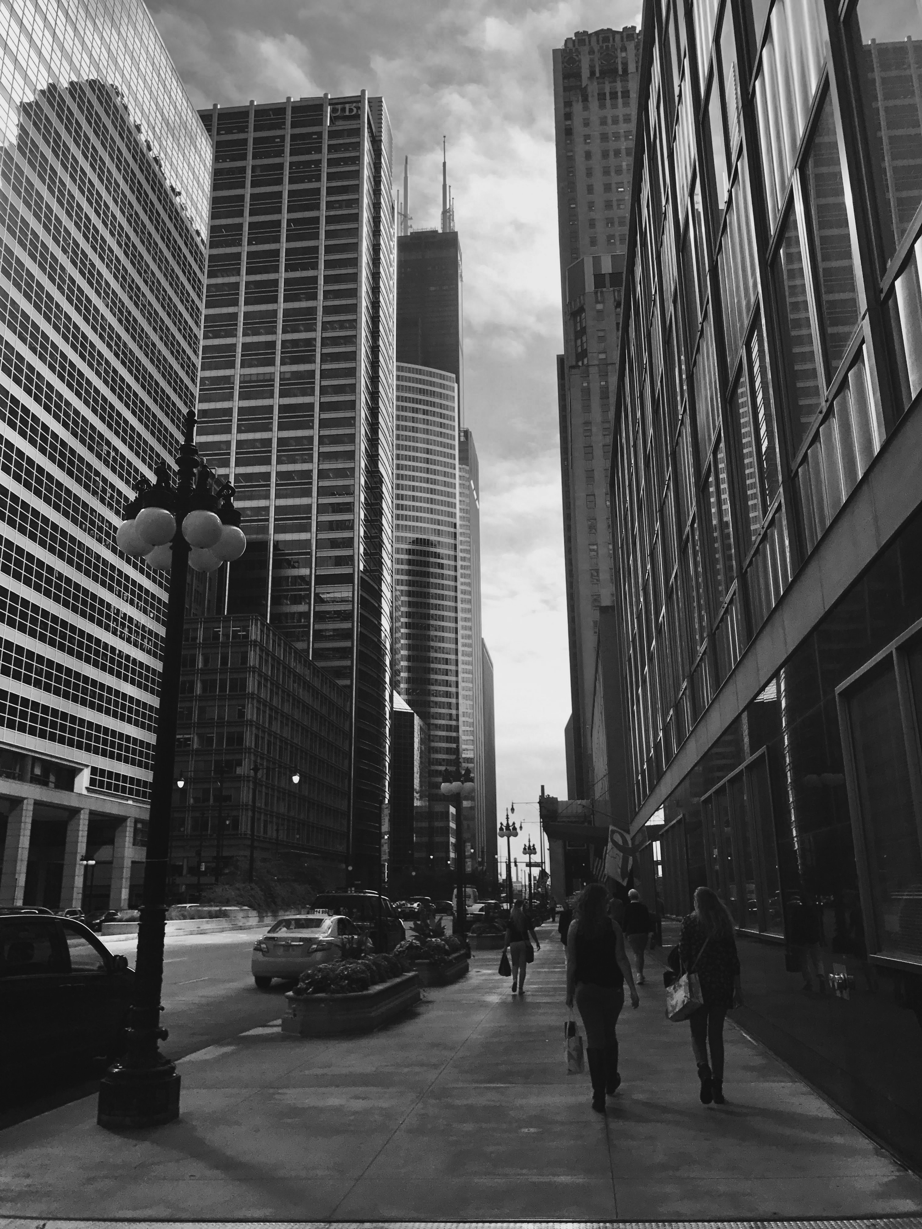 A black and white shot of the streets in chicago, looking upward to see the sears tower on the left. People and cars line the road that extends into the distance. 
