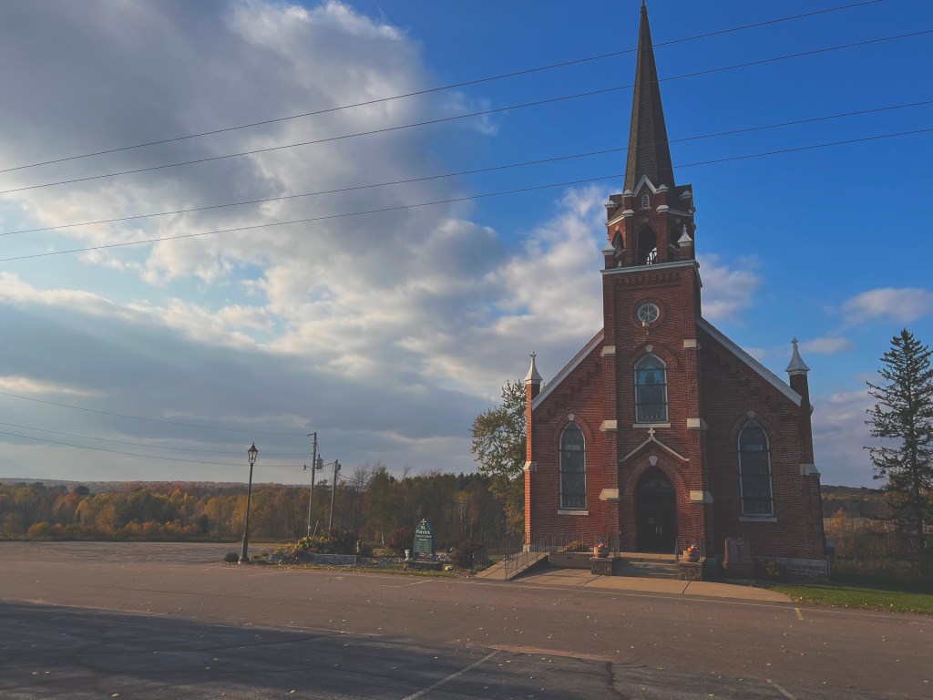 A softly lit red brick church in the countryside. The sun illuminates the side of the church which has a singular tall steeple. In the distance rolling hills burst with fall foliage.