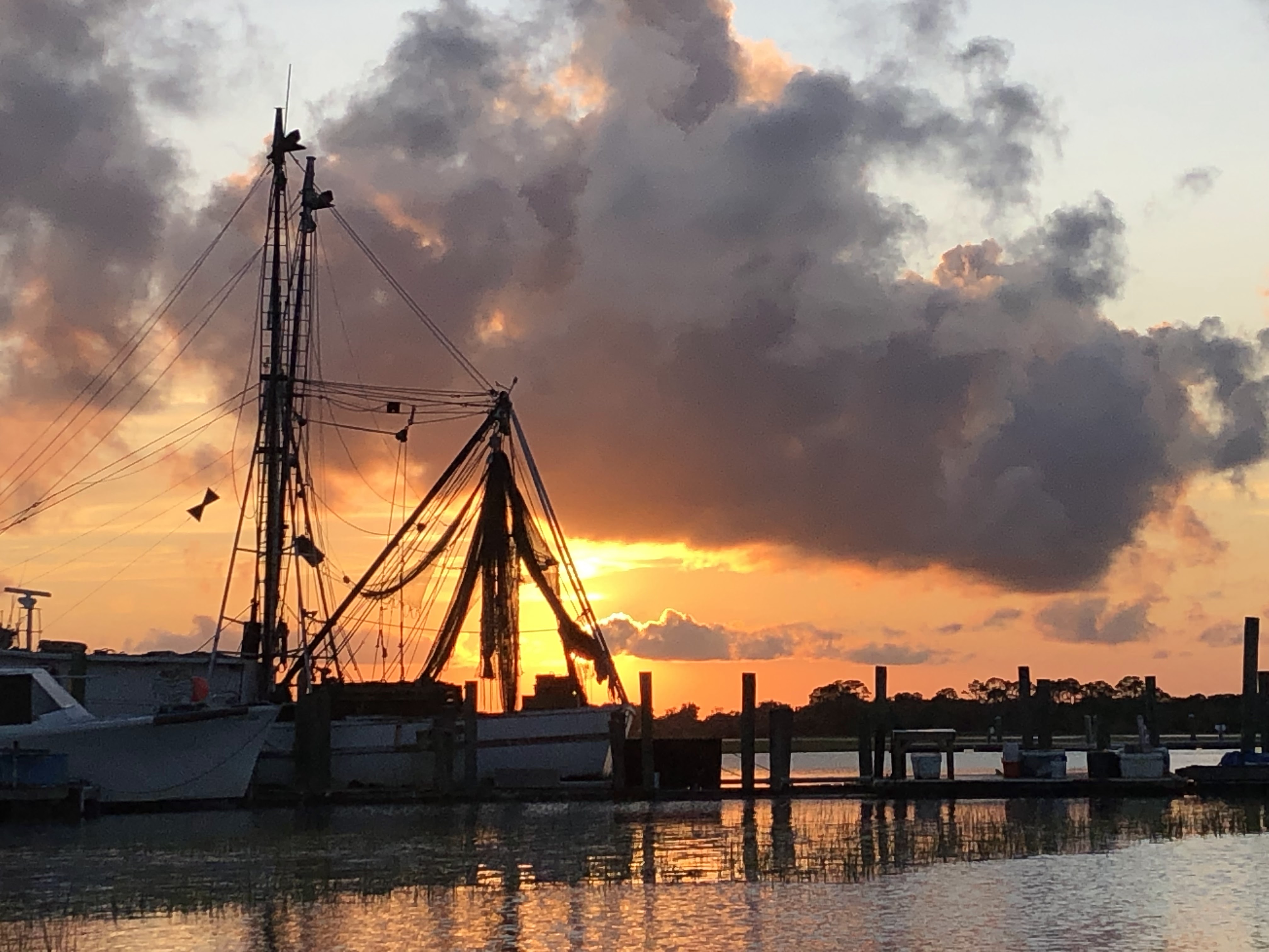 A vibrant orange sun sets, lowering into the frame of the large fishing boat that is docked. A large puffy cloud hangs above, the water rippling gently below.
