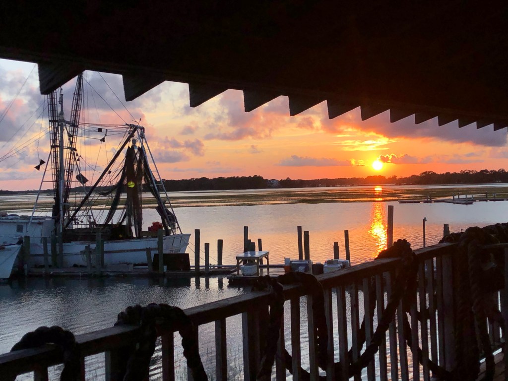 Through the silhouette of eaves and a balcony railing draped with thick fishing ropes you can see the fishing boat and a vibrant sunset in the distance. The bright orange sun sets over the marsh with pink and lavender clouds drifting overhead.