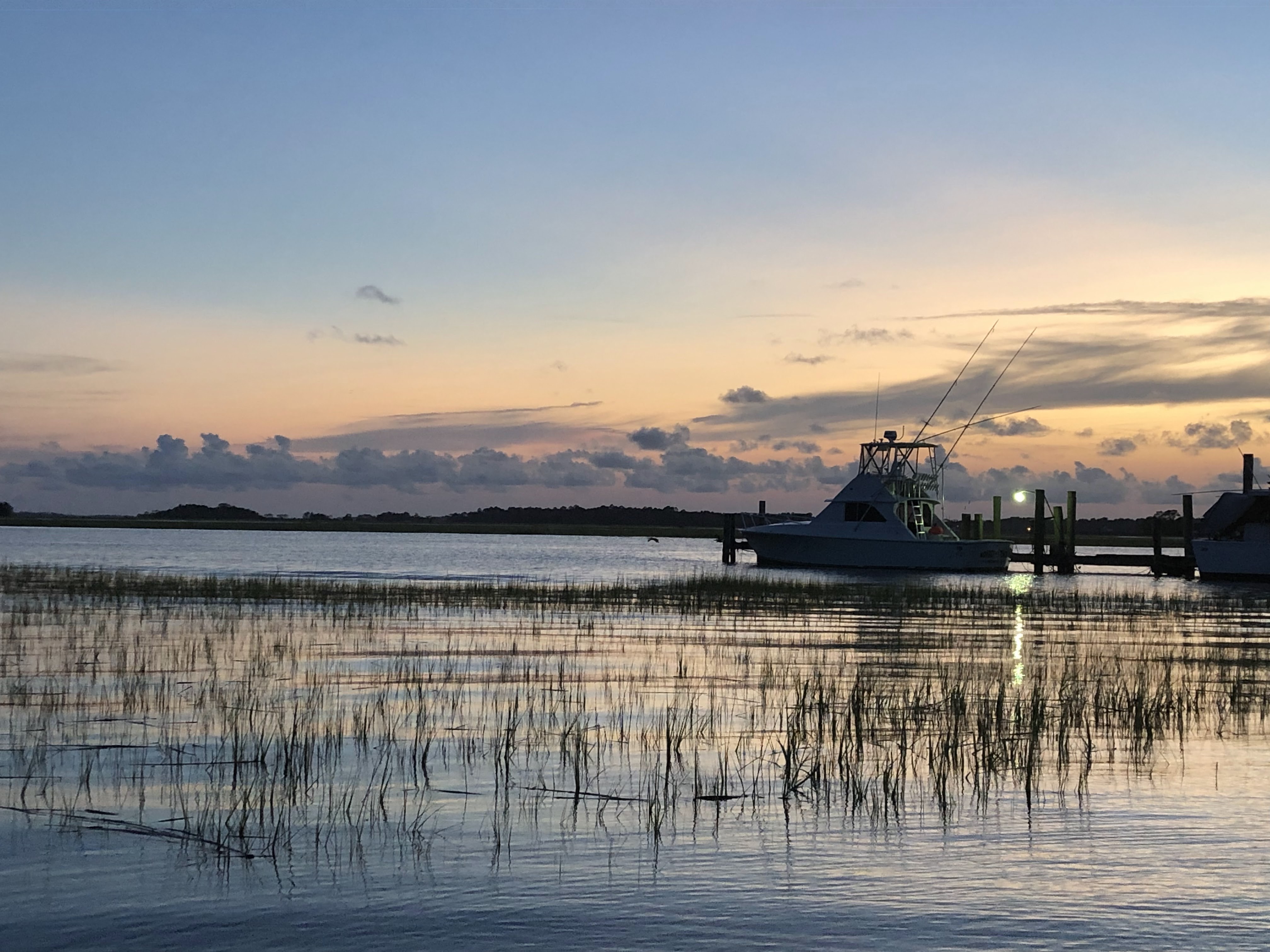 Close up on a small fishing boat at the end of the docks, the grassy marshland meets the inlet. The mainland in the distance is darkened, meeting a dark lavender cloudy line that fades to soft orange. 