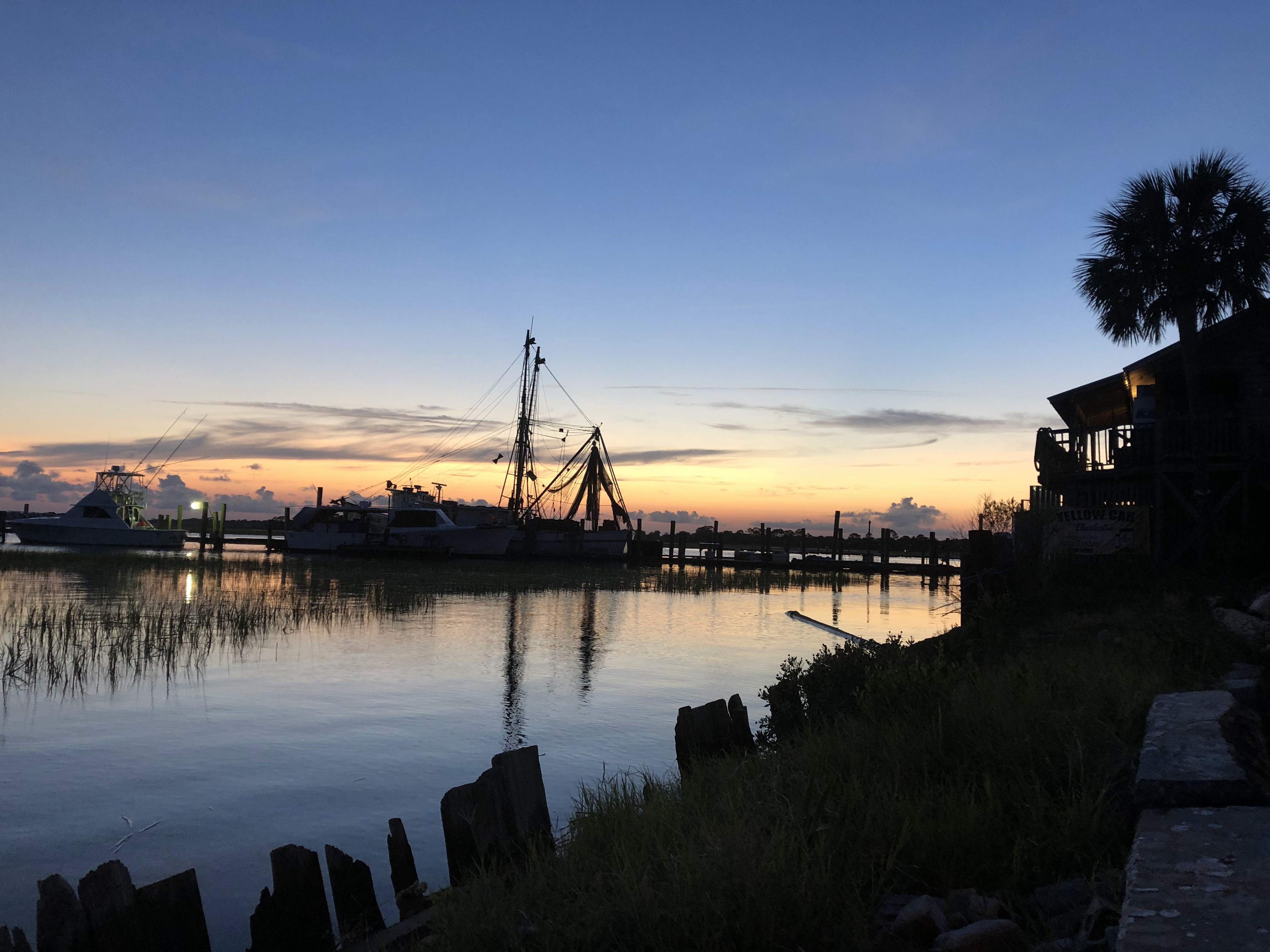 Drifting into the blue hour now, the entire scene a silhouette. The fishing boat sits centrally, the pier connecting it to the eatery where a tall palm tree hangs abovehead. The choppy plank board fence lining the water extends to meet us. The sky mostly clear now with trails of clouds hanging above a soft orange glow on the horizon.