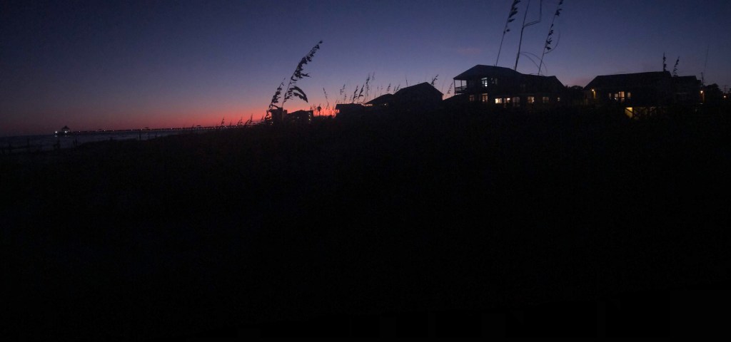 A purple to pink sunset fades along the shoreline. Tall grasses sway making way to silhouette's of beach houses overlooking the ocean. 