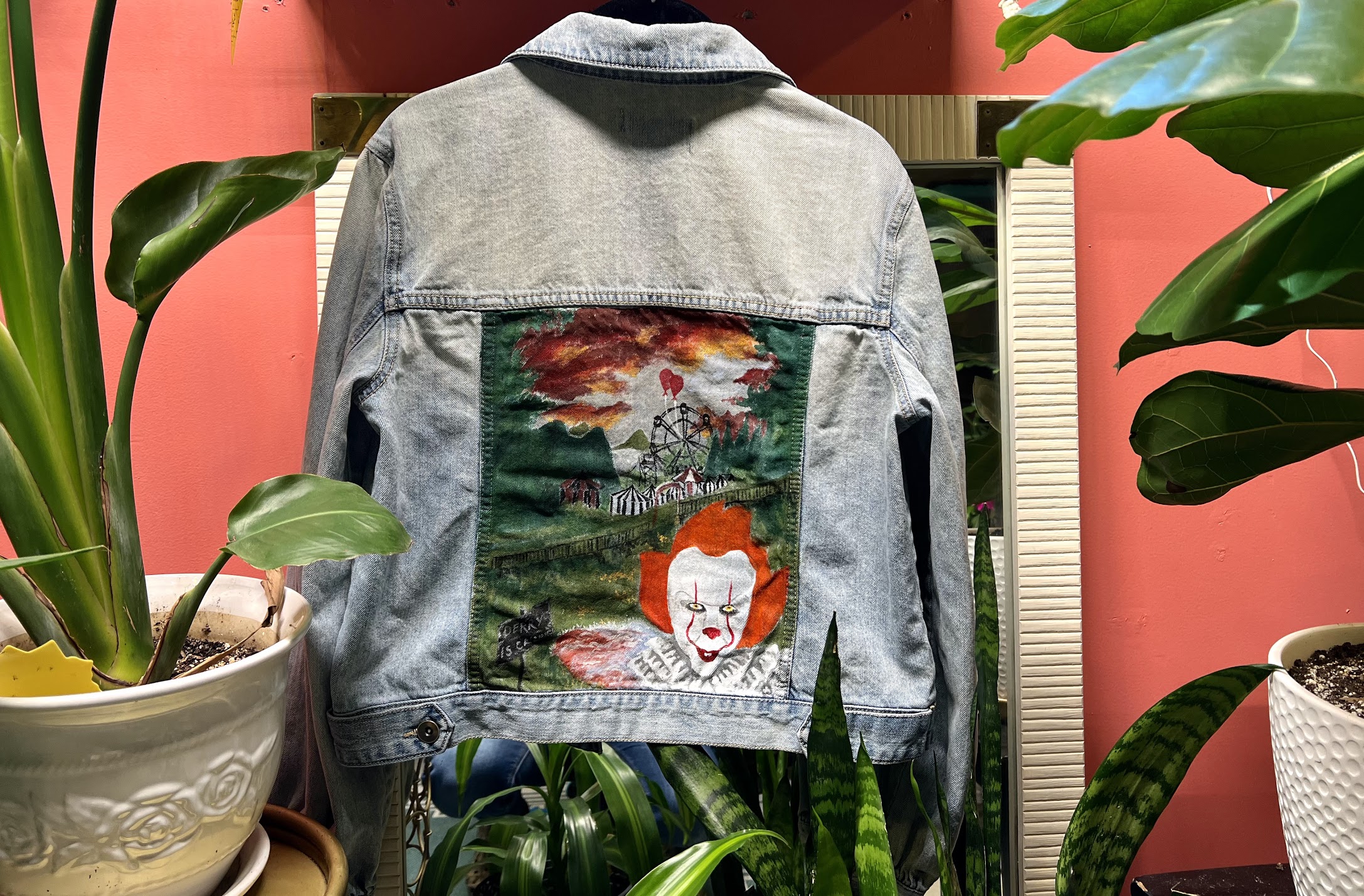 A light wash denim jacket, in the large rectangular back panel is a painting looking into the fairgrounds of Derry from the remake of IT. Trees surround the fair grounds with a ferris wheel in the center with two red balloons flying up from it to meet the red and orange hued clouds. In the foreground on the right is the new version of Pennywise the clown.