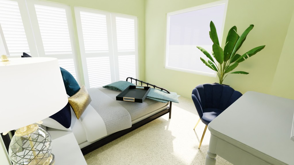 View of the bedroom as you enter the room, the walls are a soft moss color and the bed is covered in deep blue and gold pillows. There’s a white vanity with a teal scalloped velvet chair on the wall to the right and a large fake banana tree against the window that is directly across from the bed as you enter.