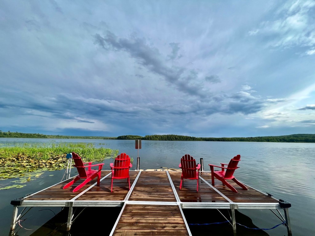 a wooden T shaped pier flanked with 4 red Adirondack chairs stand prominently over a calm lake. The dock is wet. To the left, lily pads and grass reach up through the water, glowing in the sun. Above head the sky is menacing with dark storm clouds, one shaped like a sideways V moving rapidly over the lake.