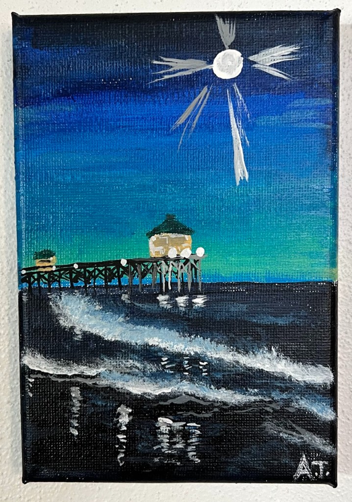 A blue night time scene with a moon rising over an illuminated pier that stretches out over the navy blue gently rolling ocean meeting the deep black sand on shore