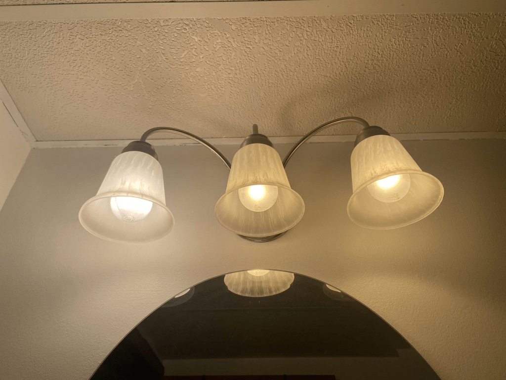 Three headed fixture against white walls the left bulb is super bright and white and lighting up the whole space. The two other bulbs look warm and faded in comparison but much gentler to look at.