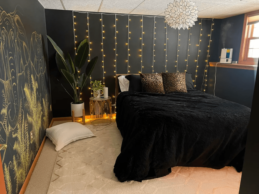 A view into my all black bedroom with a black bed on a white carpet topped with leopard pillows. The window shines bright light in on the left. Against the far wall are curtain twinkle lights. Centered over the bed is a large lotus light fixture. 
