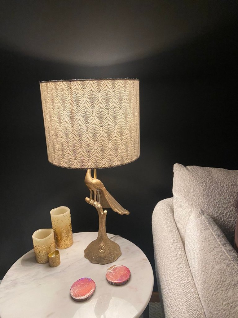 My fun gold peacock shaped lamp sitting atop a marble coffee table. The wall behind it is black and a soft glow radiates illuminating this corner of my couch.