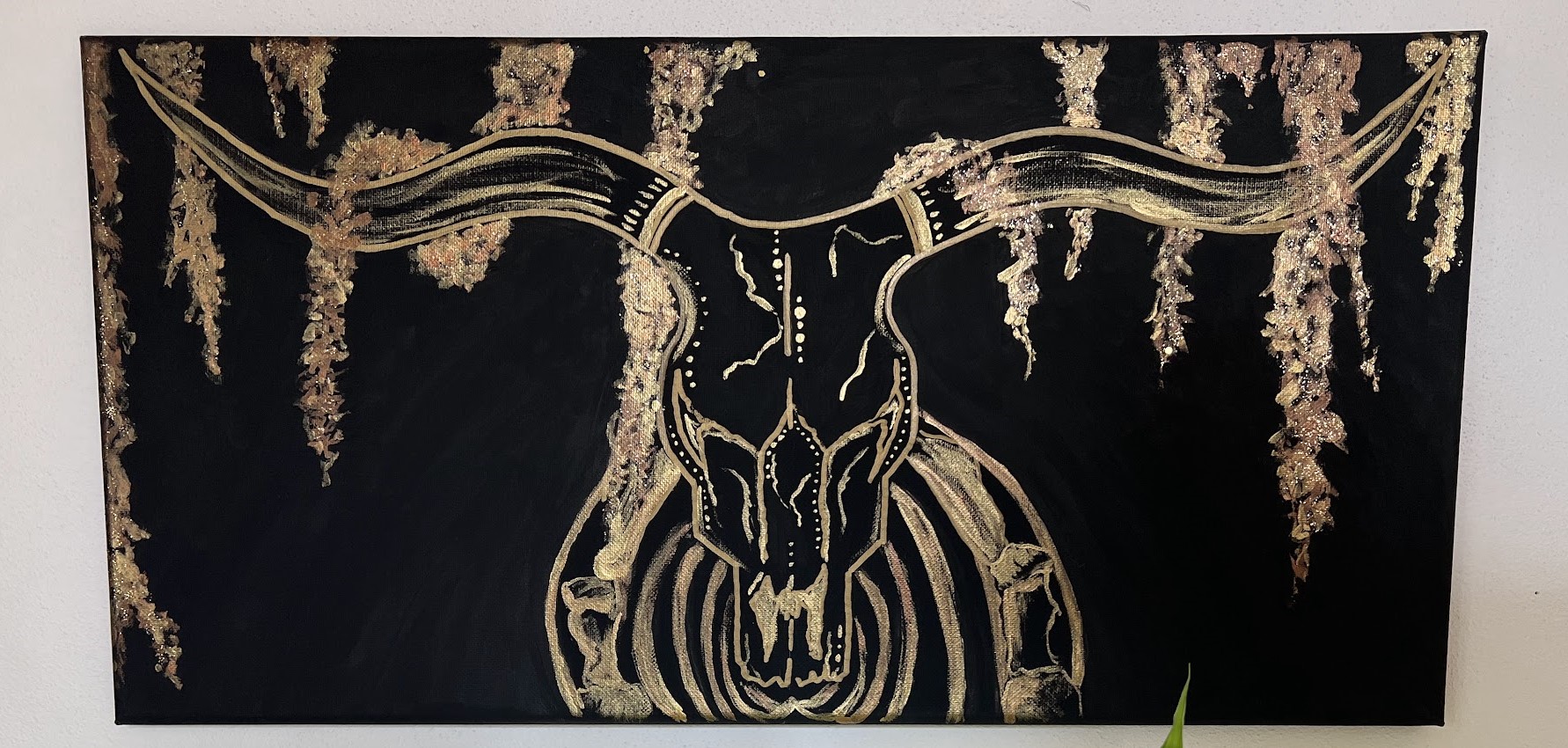 a horizontal black canvas with an outline in gold of a longhorn skeleton central on the canvas. It's horns outstretch along the top where they meet long strands of spanish moss. the skull is heavily decorated in dots