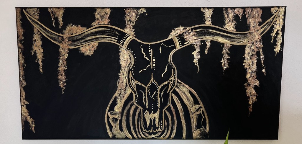 a horizontal black canvas with an outline in gold of a longhorn skeleton central on the canvas. It's horns outstretch along the top where they meet long strands of spanish moss. the skull is heavily decorated in dots