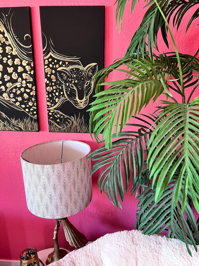 a close up of the palm in the corner with the papasan chair, it's leaves reaching slightly in front of the golden leopard triptych which glows beautifully from this angle. Its hung above a peacock lamp with a white and gold art deco lamp shade
