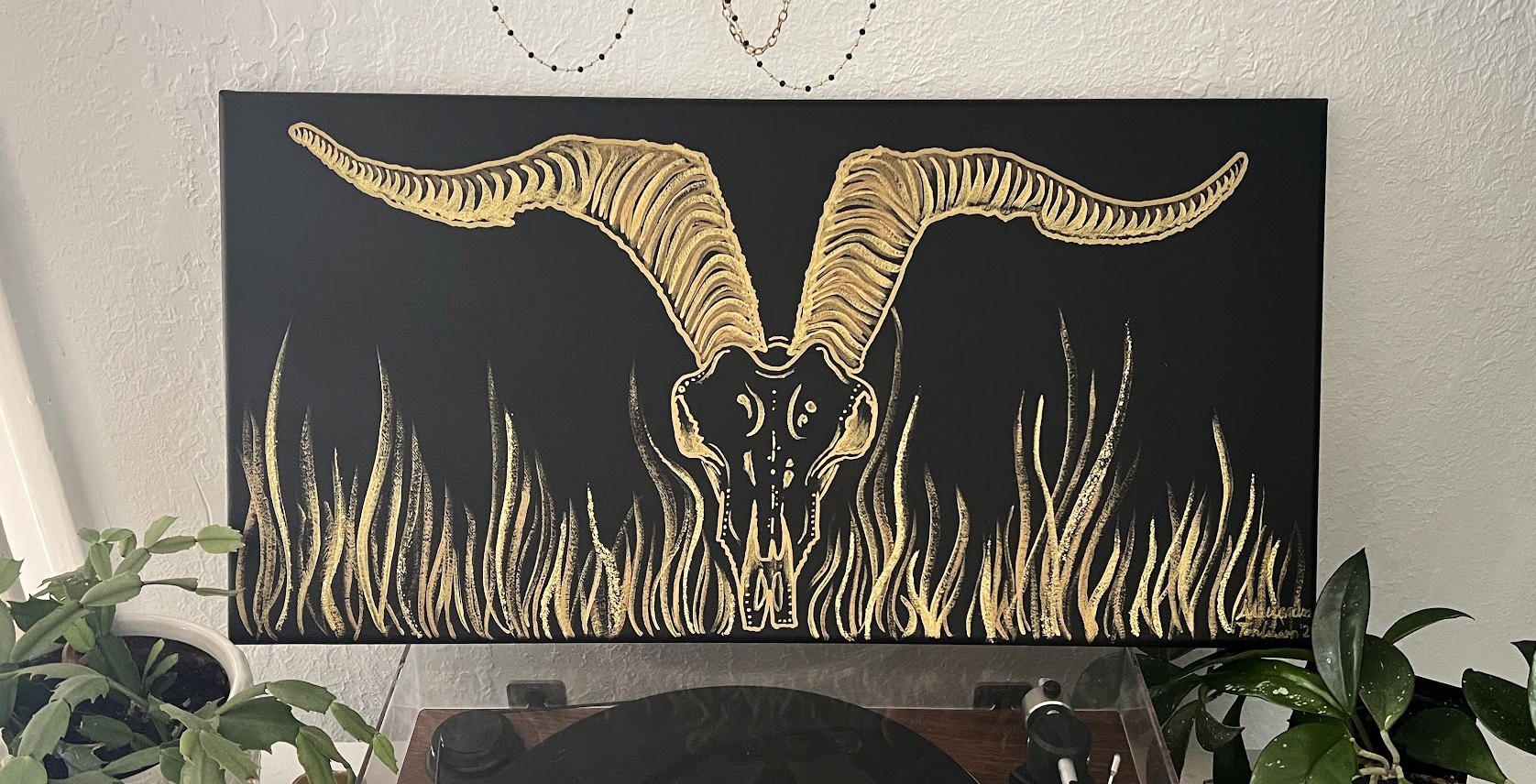 a golden goat skull with long curly horns flanged out across the top. The skull done in gold, menacing above tall fronds of grass. 