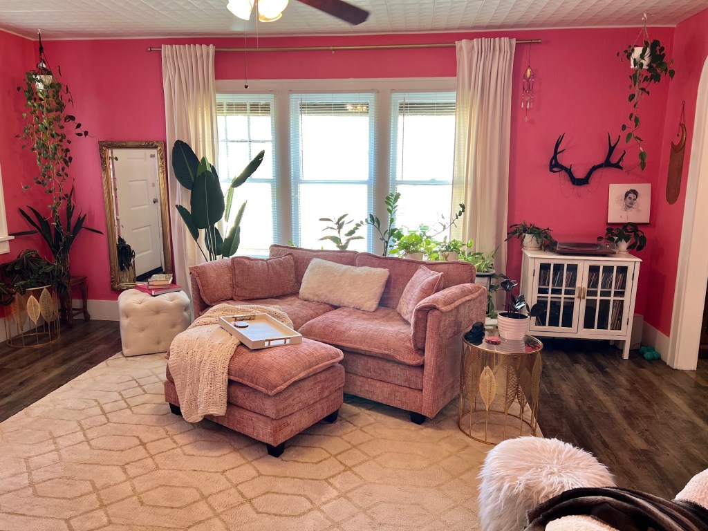 Entering the living area the walls are painted a rich watermelon pink, the ceiling, a square repeating white pattern. A soft lighter pink couch and ottoman sit angled in front of 3 windows framed by tall ivory curtains, atop a white and gold geometric rug. The couch surrounded by plants and topped with creamy accents.