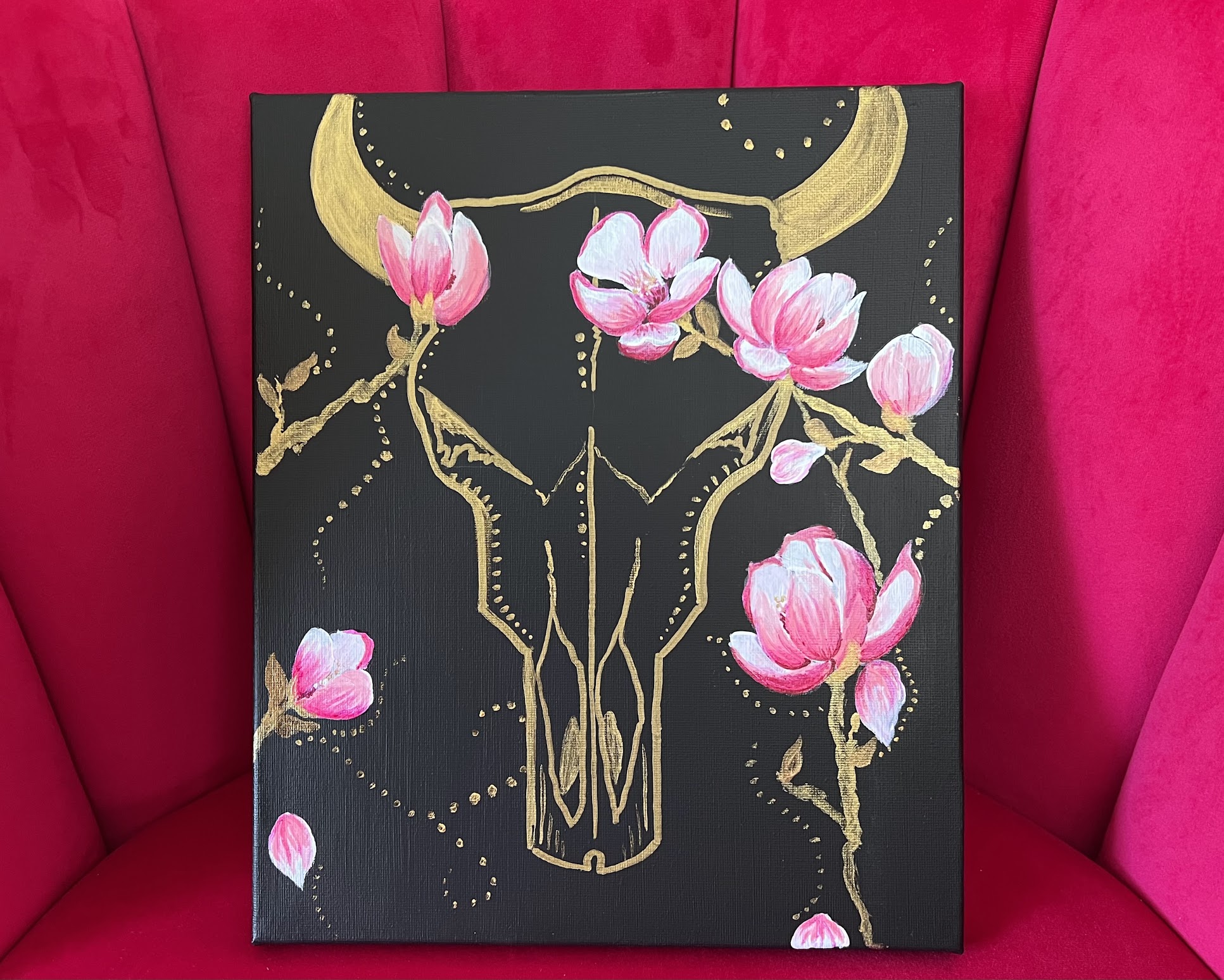 a golden cow skull on black canvas, pink magnolias extend across the top of the skull and up along the sides where some petals fall away. Swirls of golden dots fill the background