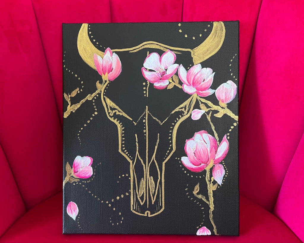 a golden cow skull on black canvas, pink magnolias extend across the top of the skull and up along the sides where some petals fall away. Swirls of golden dots fill the background