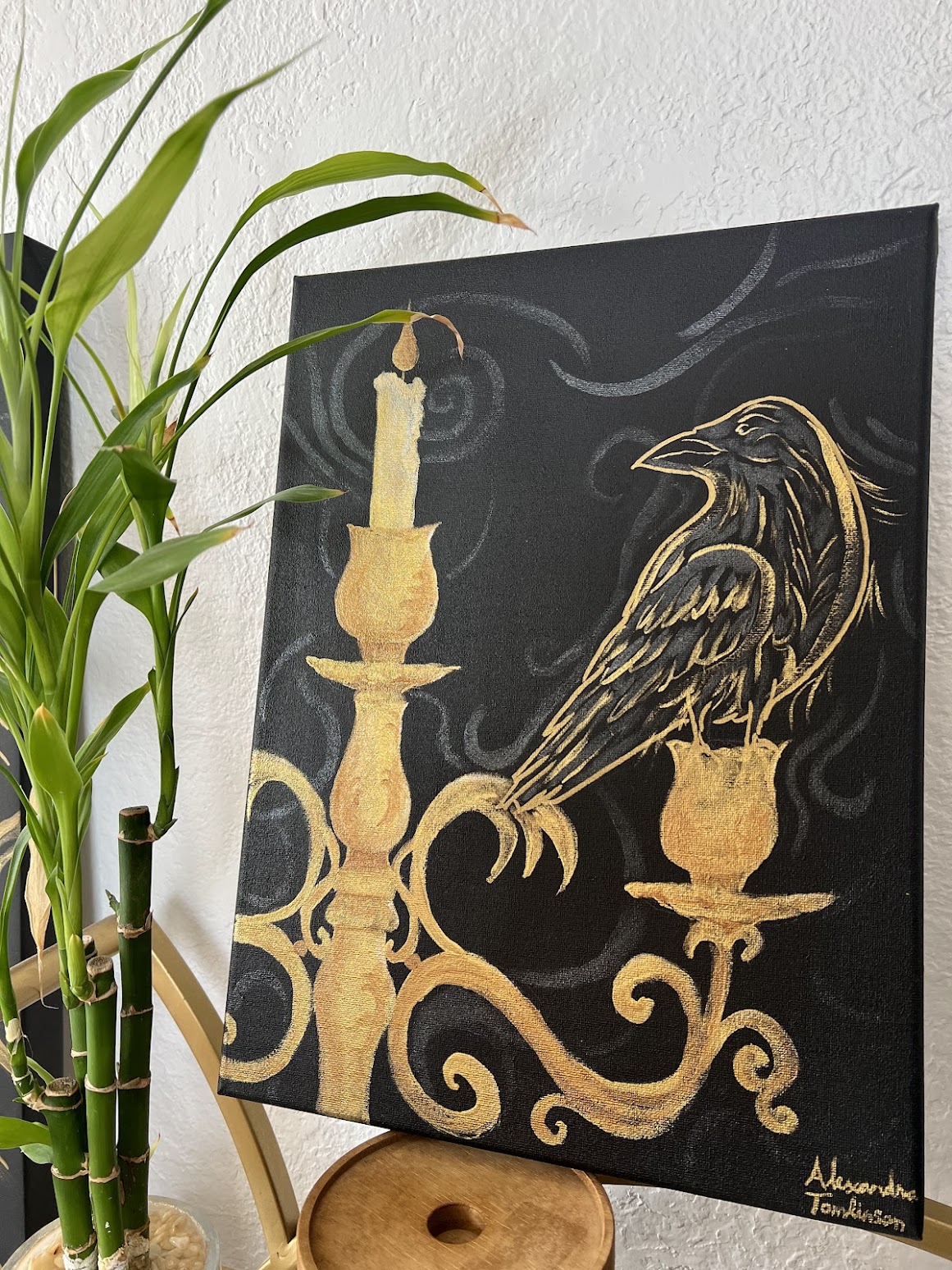 a vintage golden candelabra sits off center on the left, with a single candle burning at the top. Perched on the right arm of the candelabra is a large raven flocked with gold and black feathers. Swirls dance around the background in a glittering black in this edgar allen poe inspired piece