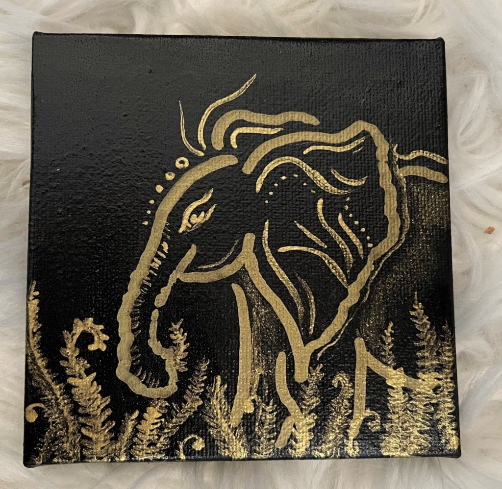 a small square shiny black canvas with a baby elephant outlined in gold accentuated with swirls and dots standing in a bed of ferns. This piece is gold and black.