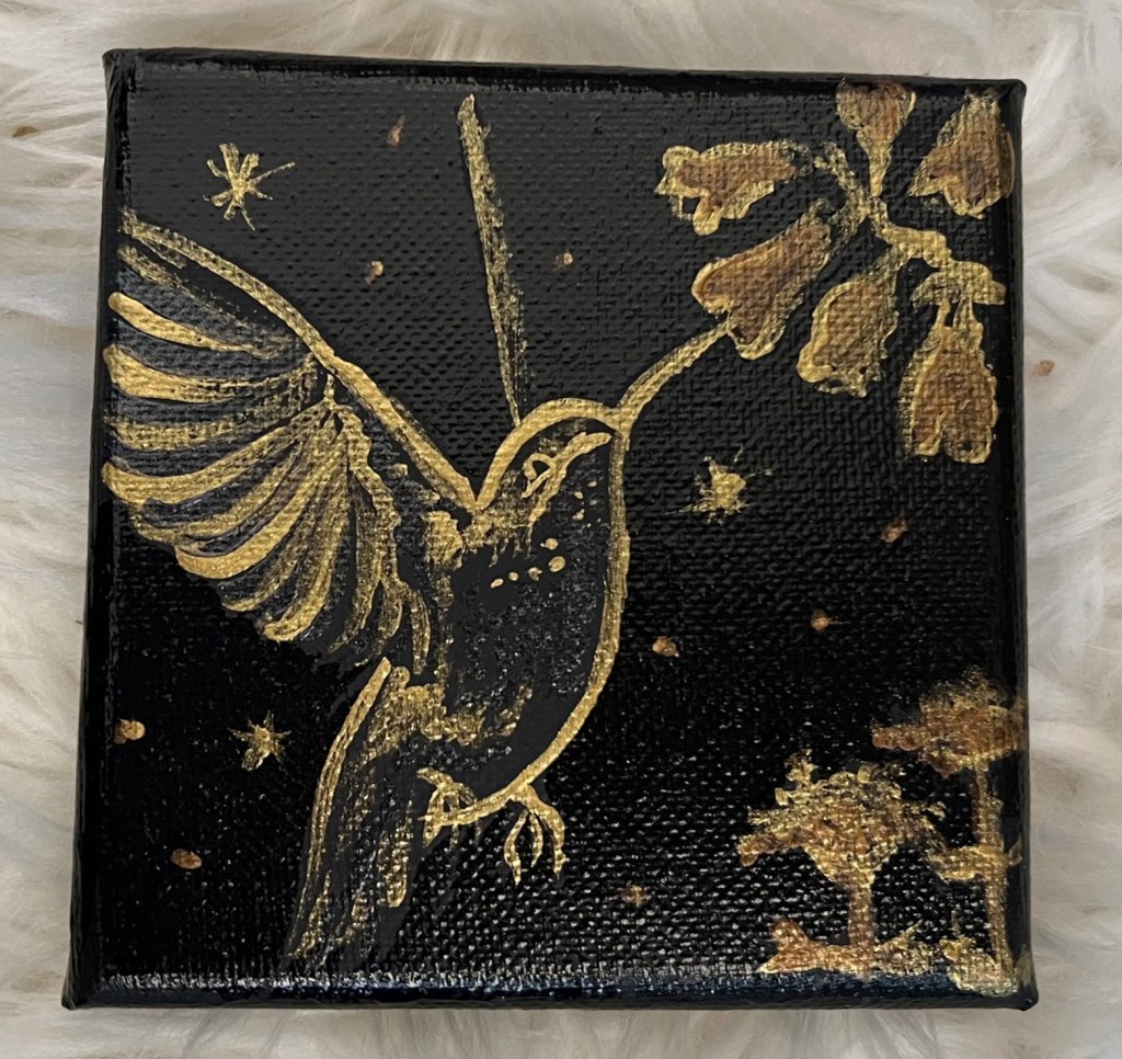 a small shiny black canvas in my classic gold and black style features a mid flight hummingbird, it's body glittering black sipping from bronze blue bell flowers, more flowers poking up below. The background dotted with gold and bronze specks meant to be fireflies and stars against the night sky.